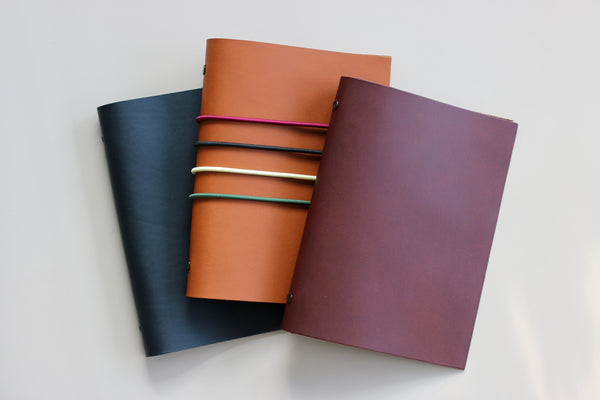 Three leather-bound notebooks in black, brown, and purple on a light gray background