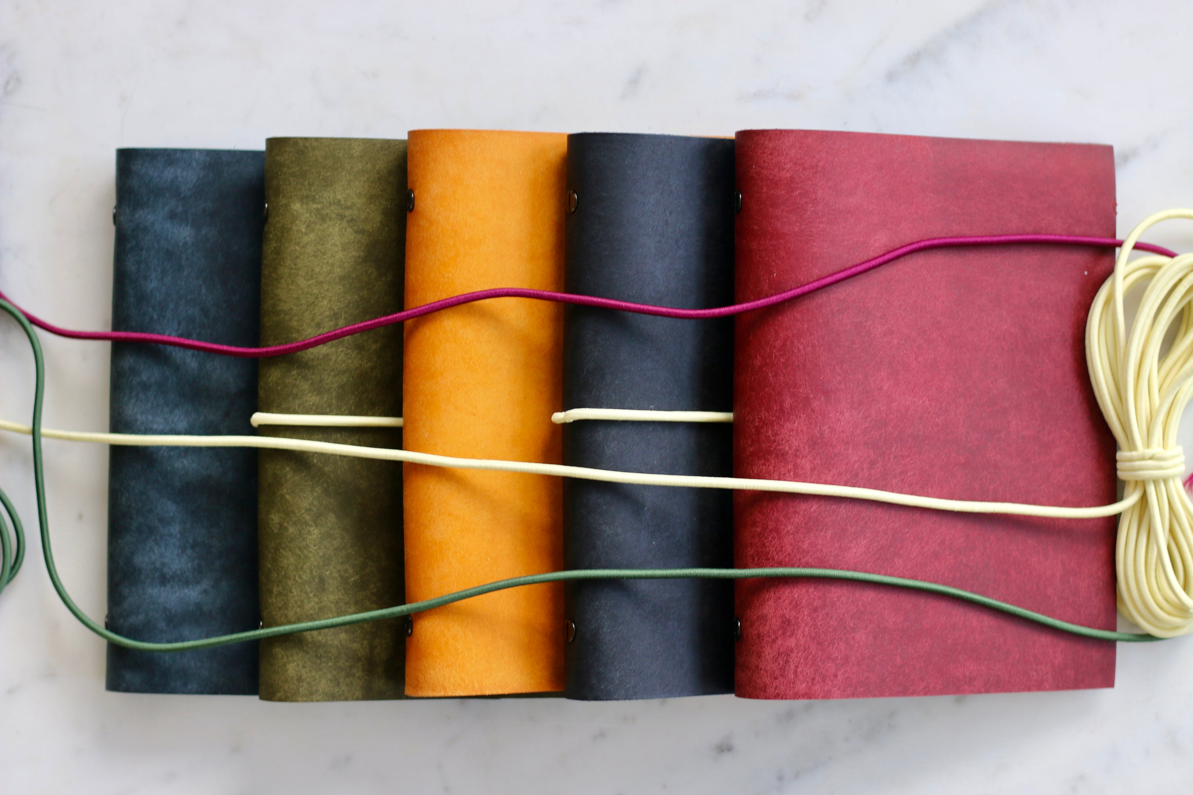 Five leather-bound books in different colors tied together with colorful strings on a marble surface.