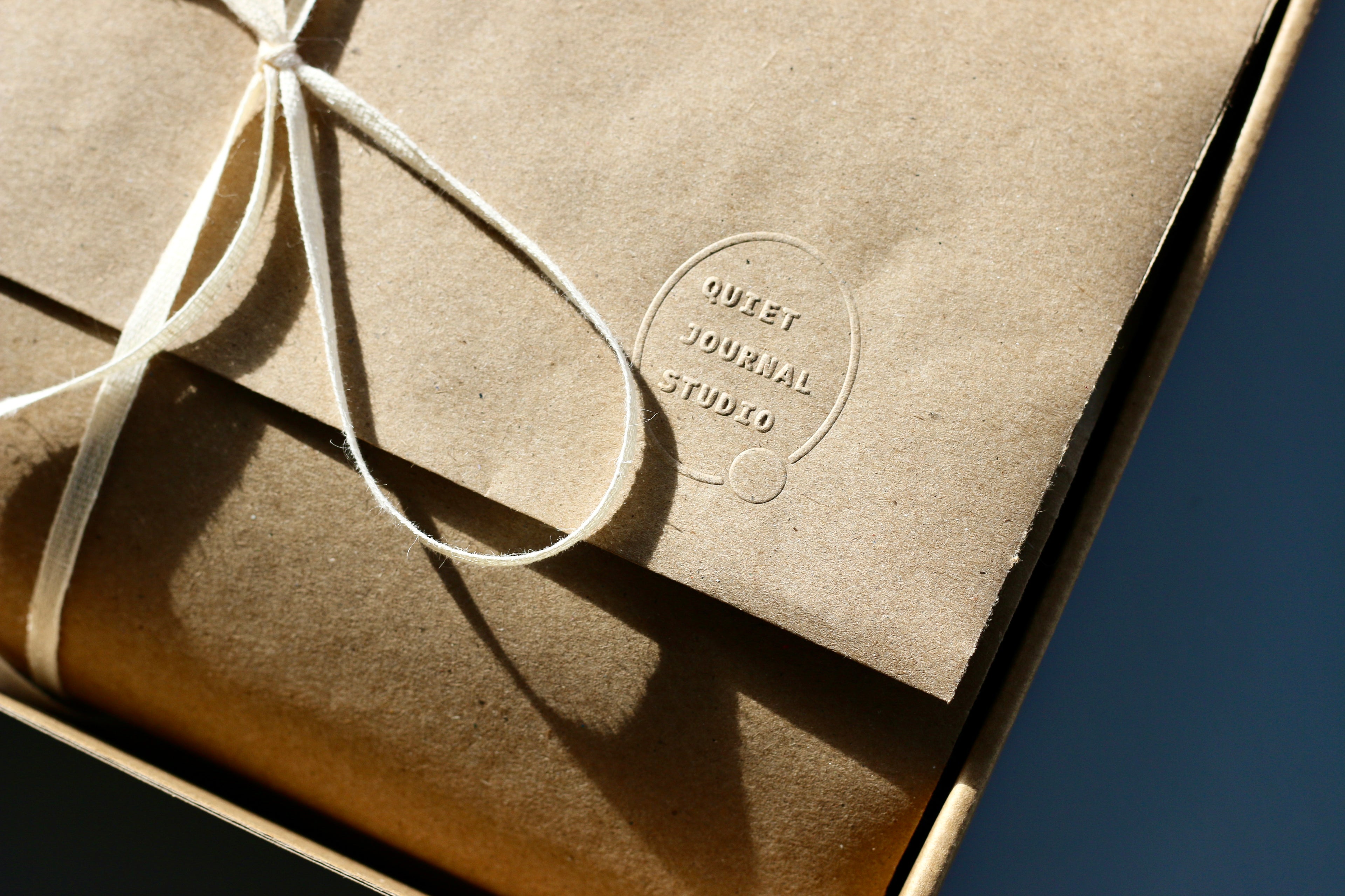 Brown paper package with a string and 'Quiet Journal Studio' logo on a dark background