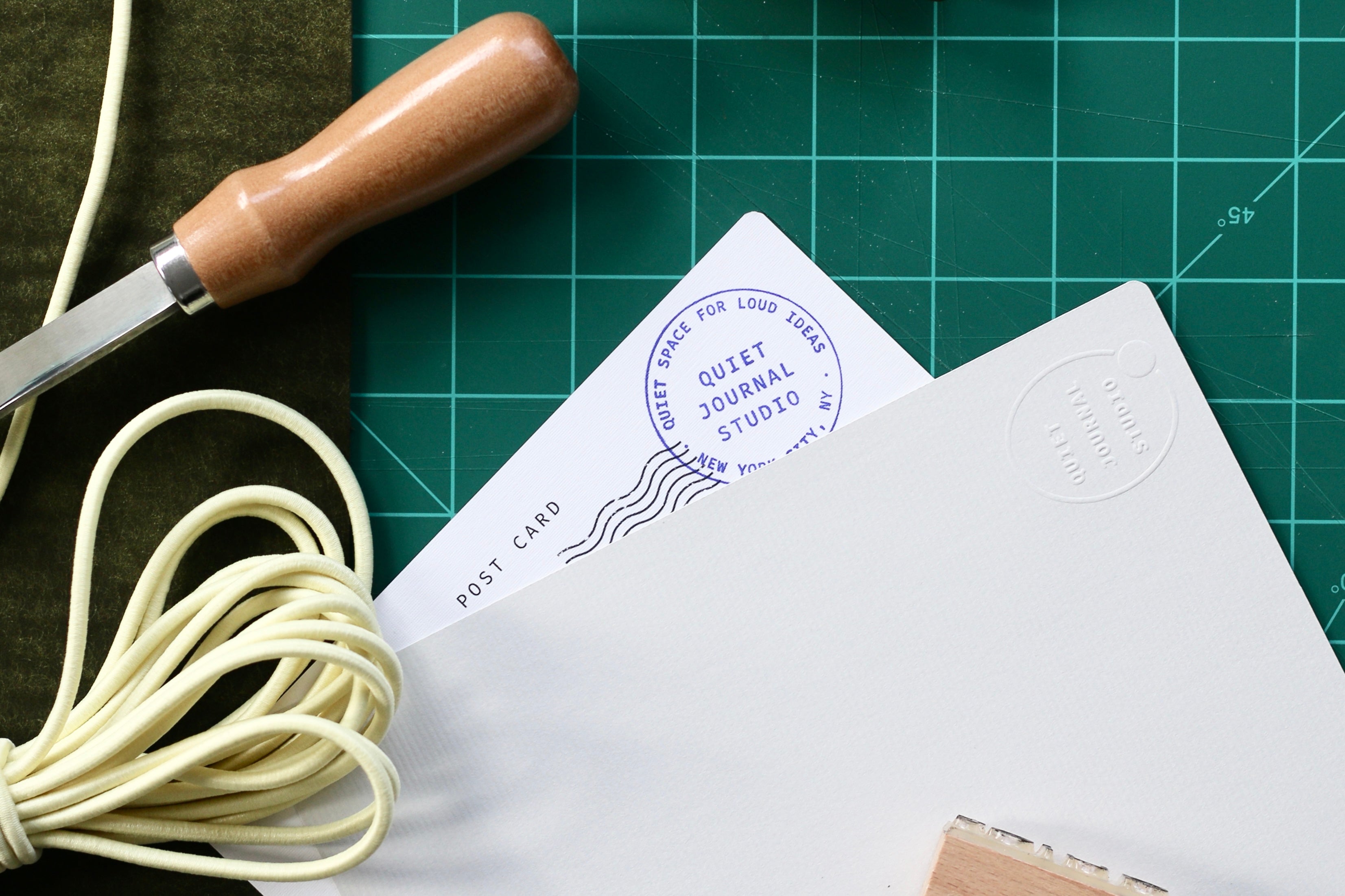 White postcard with embossed text on a green cutting mat with tools.