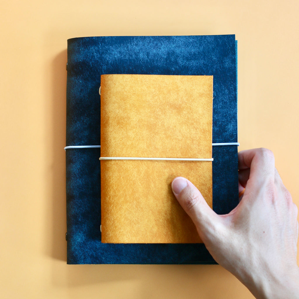 Hand holding a medium blue and a small yellow journal against a yellow background