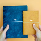 Two notebooks, one blue labeled A5 and one yellow labeled A6, held against a beige background.