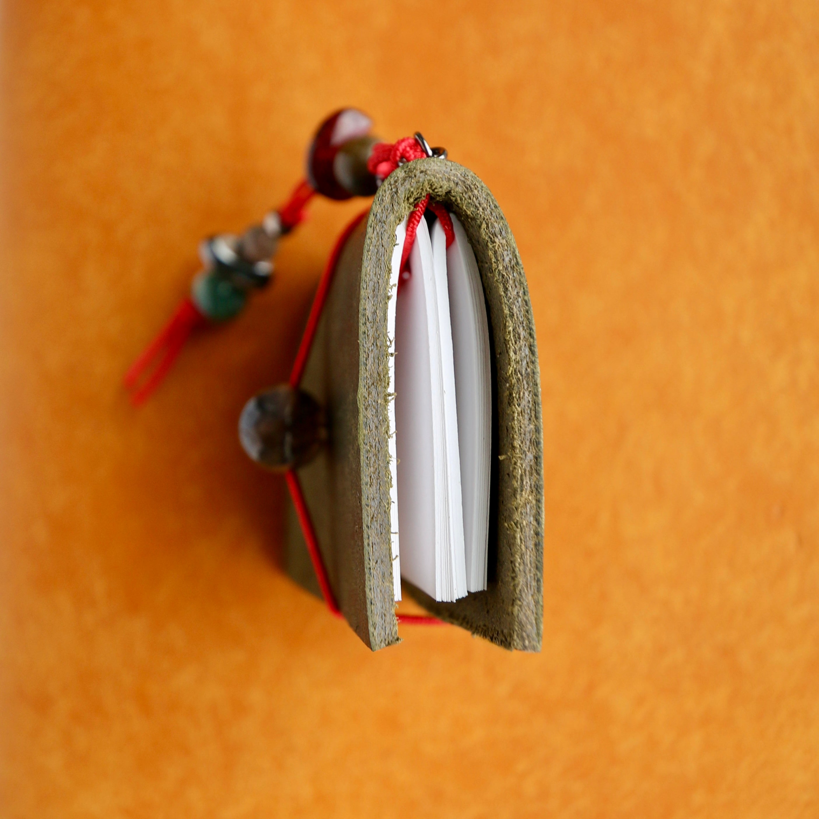 A small handmade journal with a textured olive green cover, white pages, and red cord with decorative beads, shown on a warm orange background.