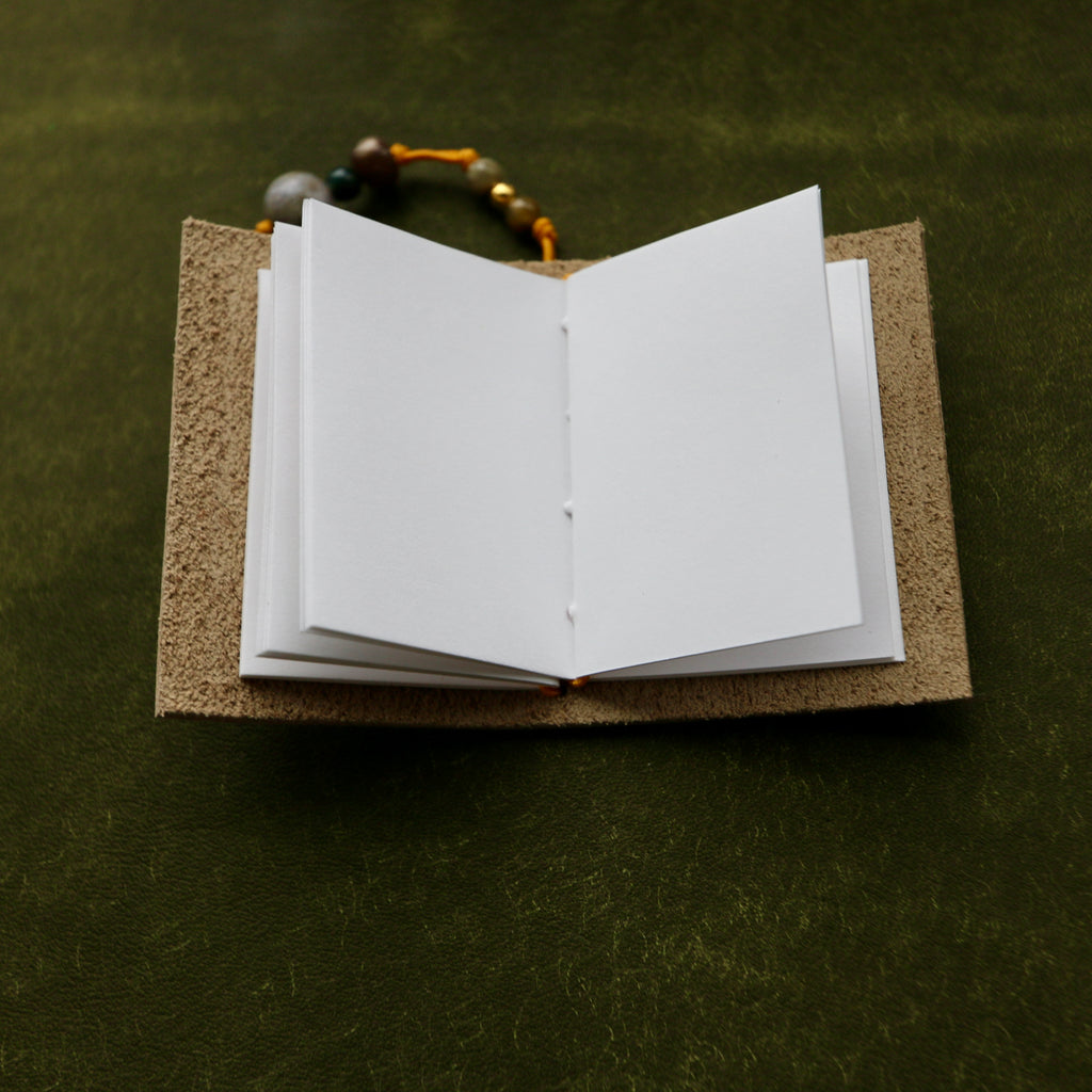 An open blank notebook with white pages and a textured brown cover, placed on a dark olive green textured surface with a beaded cord resting behind it.