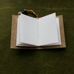 An open blank notebook with white pages and a textured brown cover, placed on a dark olive green textured surface with a beaded cord resting behind it.