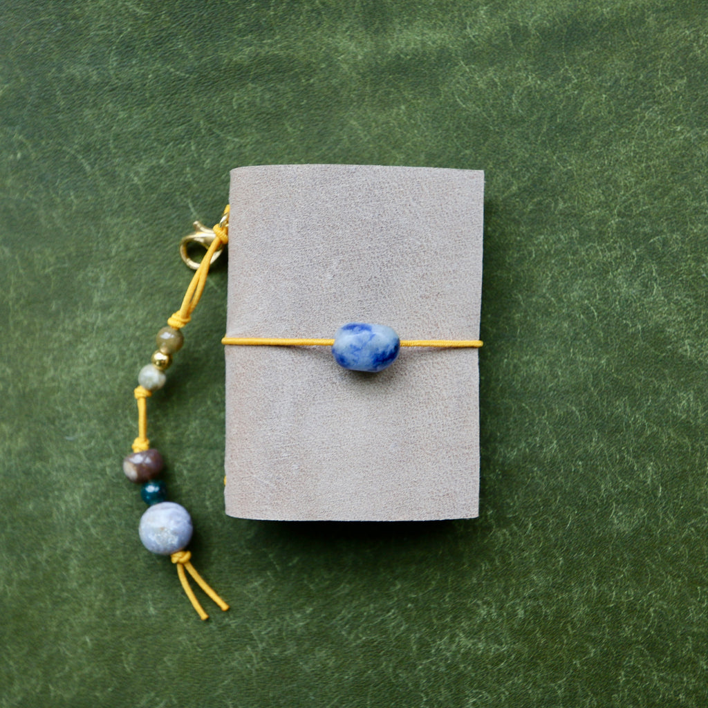 A small gray pouch or notebook tied with a yellow cord and a blue stone bead, shown on a textured green background. A matching beaded cord with blue and gray beads hangs from the side.