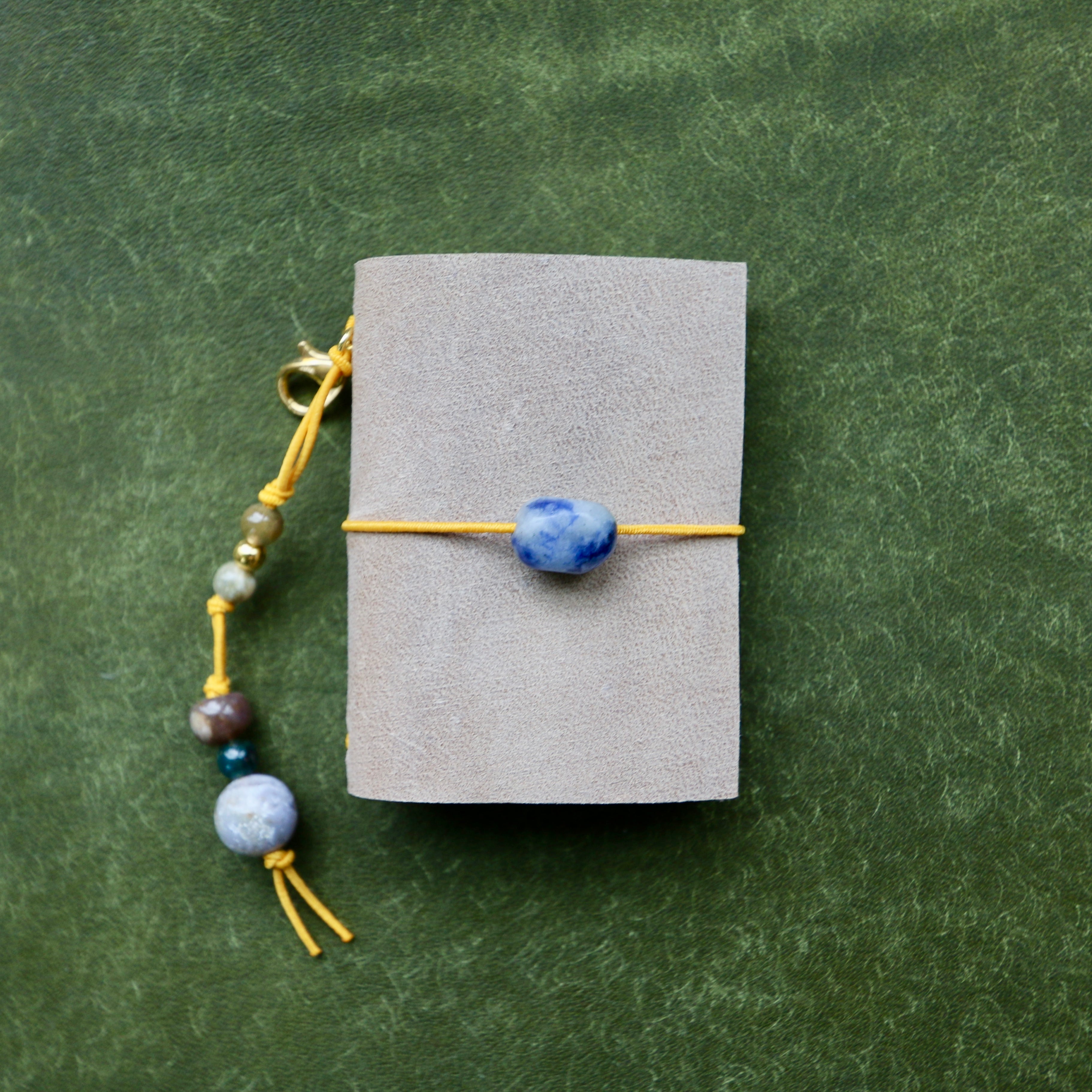 A small gray pouch or notebook tied with a yellow cord and a blue stone bead, shown on a textured green background. A matching beaded cord with blue and gray beads hangs from the side.