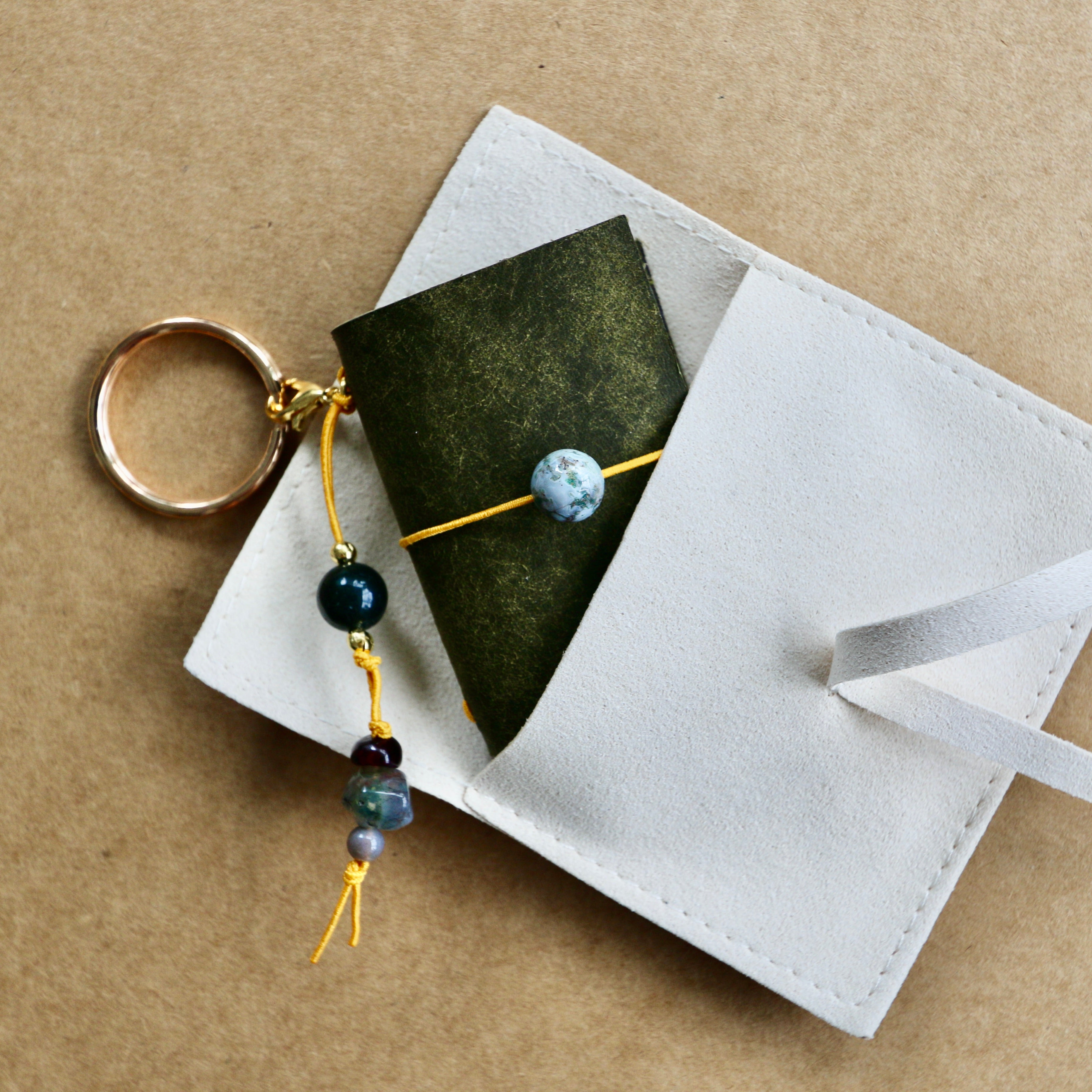 A white leather keychain holder with a gold key ring, featuring a dark green card and a white ribbon, styled on a brown kraft paper background.