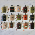 Fifteen small leather journal covers arranged in a grid on a white marble surface, each tied with a cord and bead and numbered 1 through 15. The covers come in multiple earthy colors including olive green, black, tan, brown, and terracotta.