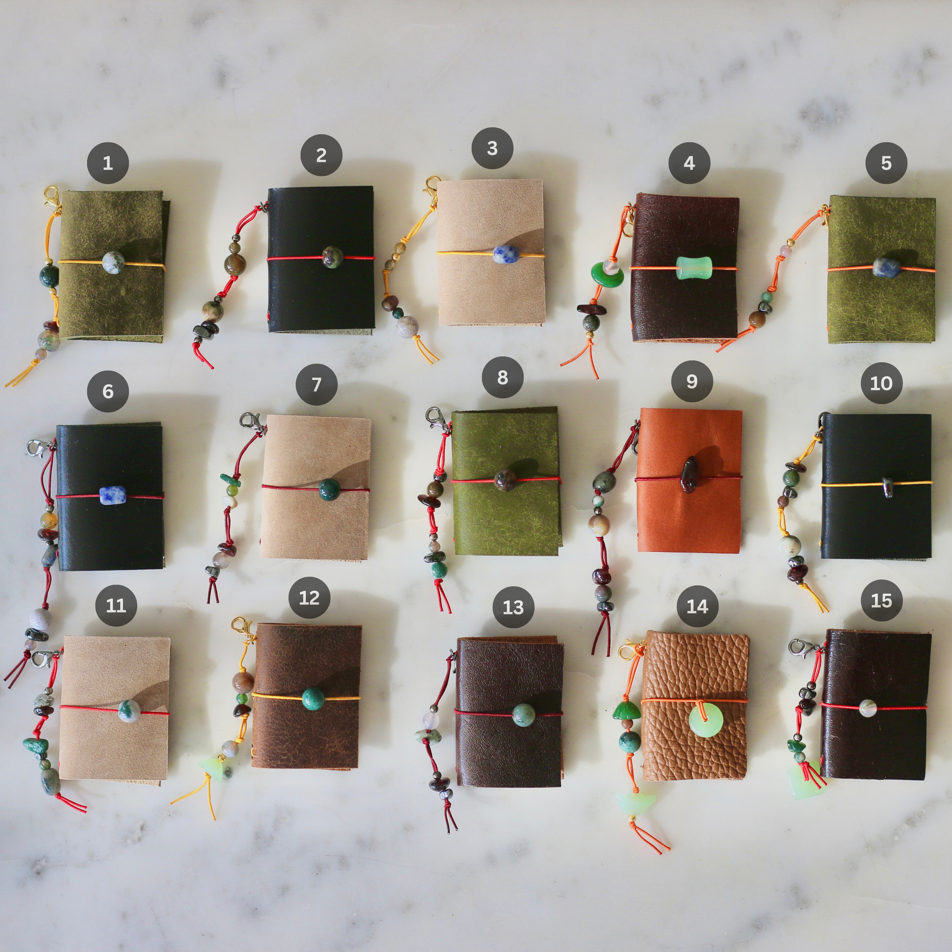 Fifteen small leather journal covers arranged in a grid on a white marble surface, each tied with a cord and bead and numbered 1 through 15. The covers come in multiple earthy colors including olive green, black, tan, brown, and terracotta.